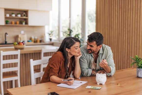 Young couple smiling while calculating expenses and planning their budget together at home, using a piggy bank and a notebook on the kitchen table, enjoying quality time