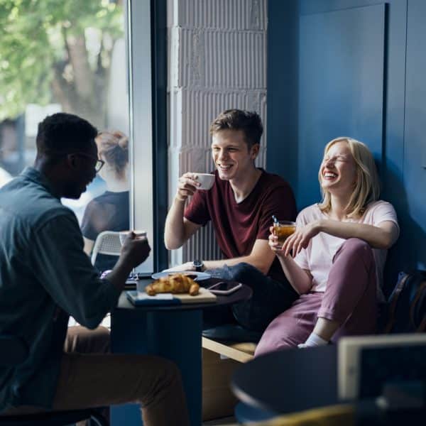 Two men and a woman talking and laughing in a coffee shop while drinking coffee and eating breakfast together