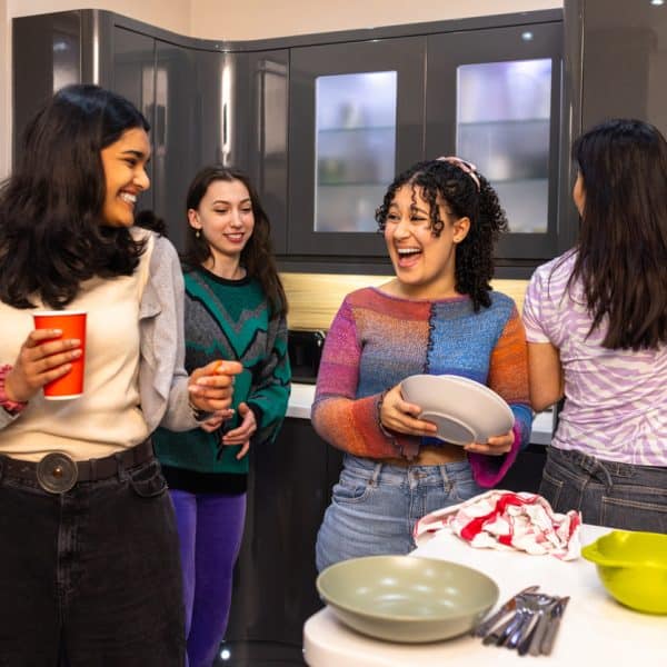 A wide shot of five diverse university students enjoying refreshments whilst cooking in a kitchen. They are talking to each other and holding red cups. They are all wearing casual clothing in a student house in Durham in the North East of England.